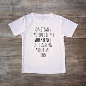 May include: White t-shirt with the text "SOMETIMES I WONDER IF MY WORKBENCH IS THINKING ABOUT ME TOO" in black. The shirt is centered on a wooden background. The brand name "Staple Stitch" is in the lower left corner.