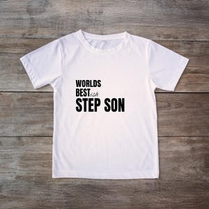 May include: A white t-shirt with the text "Worlds Best Step Son" in black. The shirt is displayed on a wooden surface.