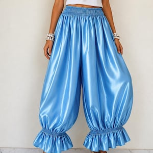 Puede incluir: Pantalones de satén azul brillante con cintura elástica alta, silueta ancha y voluminosa, y dobladillos con volantes fruncidos en los tobillos. La tela tiene un brillo suave y reflectante con un ajuste holgado y fluido en todas las piernas.