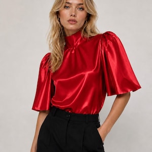 Op de afbeelding: Een felrode satijnen blouse met een hoge mock-hals en volumineuze pofmouwen tot op de elleboog. De stof heeft een gladde, reflecterende afwerking en het kledingstuk wordt gecombineerd met een effen zwarte broek met een zichtbare tailleband en knoop.