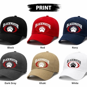 May include: Six baseball caps in black, red, navy, dark grey, khaki, and white. Each cap features the text BLACKWATER in an arch above a white bear paw print centred inside a red oval target graphic. The word PRINT appears at the top centre.