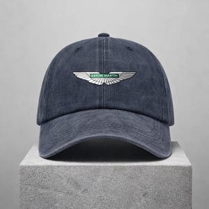 Op de afbeelding: Een marineblauwe baseballcap met het Aston Martin-logo op de voorkant. Het logo heeft zilveren vleugels en de merknaam in groen. De cap is gemaakt van een zachte, getextureerde stof.