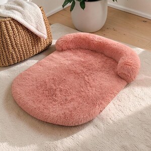 May include: A plush, dusty rose-colored pet bed with a soft, shaggy faux fur texture. The bed features a rounded, flat base and a raised, cylindrical bolster along the back edge, providing a cushioned support area for a small animal to rest.