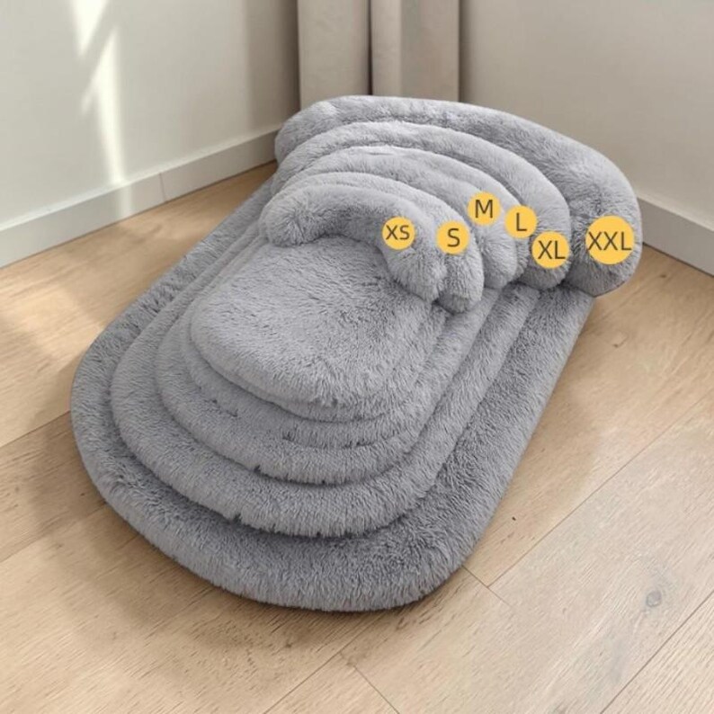 May include: A series of nested, light gray, plush, rectangular pet beds with raised edges. The beds are stacked from smallest to largest, with yellow circular labels indicating sizes XS, S, M, L, XL, and XXL placed on the raised bolster edges.