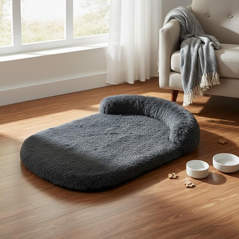 May include: A rectangular dark gray pet bed with a plush, shaggy texture and a raised bolster along one end. The bed is accompanied by two plain white ceramic bowls and several small wooden paw-shaped decorative accents on the floor.