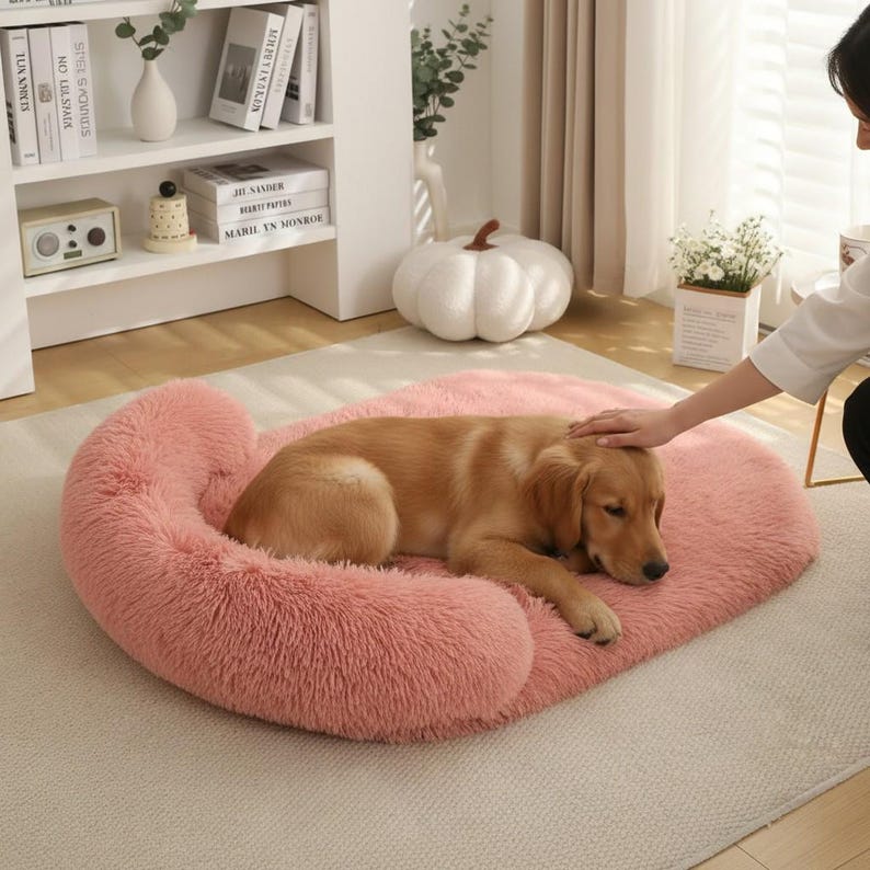 May include: A plush, oval-shaped pet bed in a soft pink color with a raised, cushioned bolster edge. The bed features a long-pile, faux fur texture and is designed for a pet to rest comfortably on its soft, padded surface.