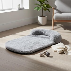 May include: A light gray, rectangular pet bed with a plush, fuzzy texture and a rounded, elevated bolster along one end. The bed is accompanied by two small, knotted rope toys and a folded, cream-colored fabric item resting nearby on the floor.
