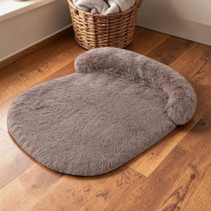 May include: A taupe-colored pet bed with a plush, faux-fur texture. The bed features a rounded, flat base and a raised, cushioned bolster along the back edge, providing a soft surface for resting. The material is uniform in color and texture.