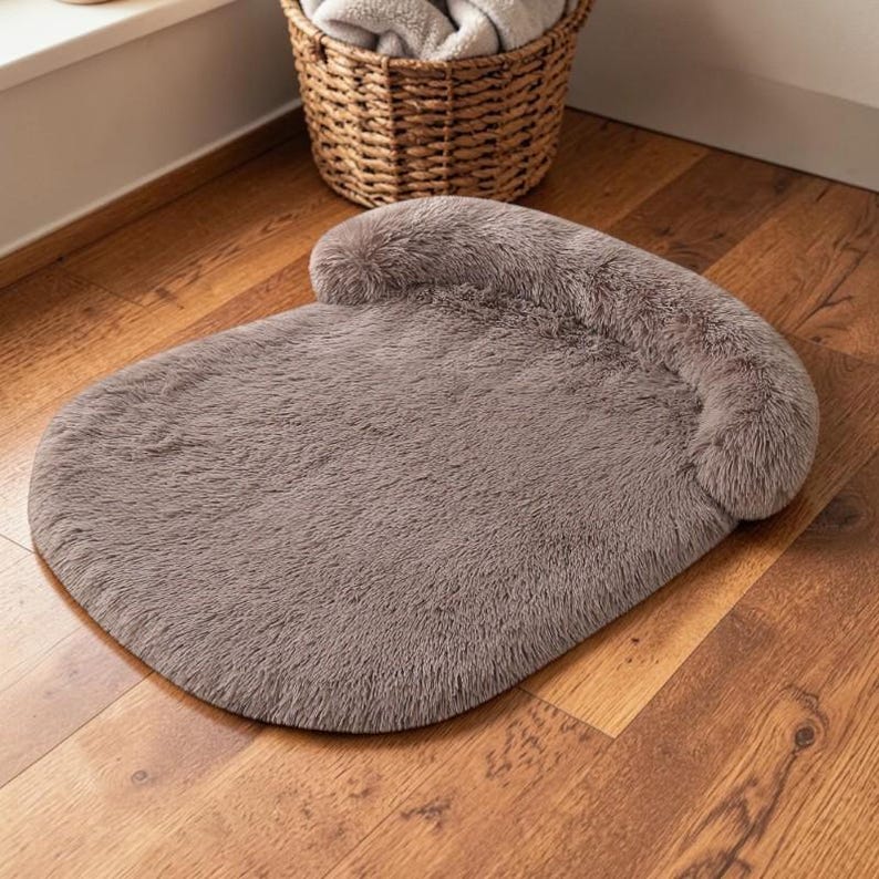 May include: A taupe-colored pet bed with a plush, faux-fur texture. The bed features a rounded, flat base and a raised, cushioned bolster along the back edge, providing a soft surface for resting. The material is uniform in color and texture.