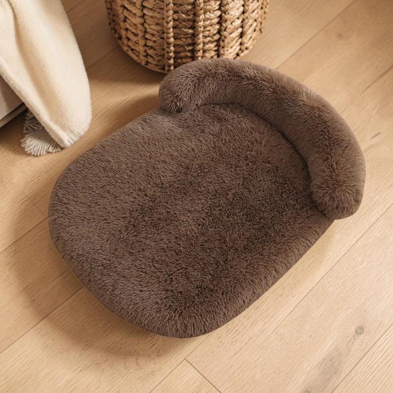 May include: A rectangular pet bed featuring a soft, dark brown faux fur texture and a raised, curved bolster along one side. The bed has a plush, cushioned surface designed for resting, with a consistent deep brown color throughout the fabric.