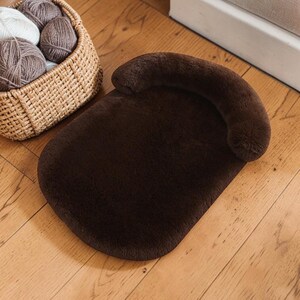 May include: A dark brown plush pet bed with a rounded rectangular base and a curved, raised bolster along one end. Beside the bed sits a woven wicker basket containing several balls of brown and white yarn, showcasing a soft, fuzzy texture.