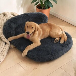 May include: A dark gray, plush, oval-shaped pet bed with raised bolster sides. A golden-colored dog with its tongue out is lying down on the soft, furry surface of the bed, which features a thick, cushioned base for comfort and support.