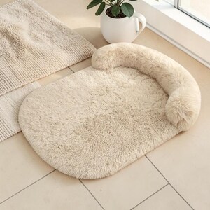 May include: An oval-shaped pet bed made of soft, cream-colored faux fur. The bed features a raised, cushioned bolster along one side, providing a comfortable resting area with a plush, shaggy texture throughout the entire surface.