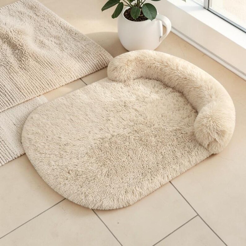 May include: An oval-shaped pet bed made of soft, cream-colored faux fur. The bed features a raised, cushioned bolster along one side, providing a comfortable resting area with a plush, shaggy texture throughout the entire surface.