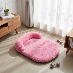 May include: A plush, oval-shaped pet bed in a solid light pink color, featuring a raised, cushioned bolster edge along one side for support. Two small, textured, multi-colored rope toys are positioned on the floor near the pet bed.