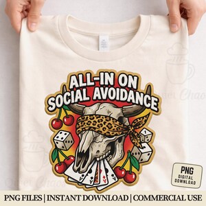 May include: Cream-colored t-shirt featuring a graphic with the text "ALL-IN ON SOCIAL AVOIDANCE". The graphic includes a skull wearing a leopard print blindfold, playing cards, dice, and cherries. The shirt is held up by two hands.
