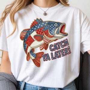 以下が含まれることがあります： 赤、白、青の愛国的な魚のデザインが施された白いTシャツ。魚は星とストライプのパッチワークデザインです。「CATCH YA LATER!」の文字は、白い星が入った青色です。