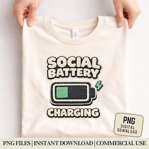 May include: Cream-colored t-shirt with the words "SOCIAL BATTERY CHARGING" above a battery icon. The battery is black with a green fill and a green lightning bolt. The shirt is held up by two hands.