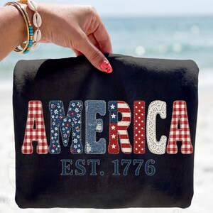 May include: Black sweatshirt with the word "AMERICA" in a patchwork design. The letters feature floral, denim, dotted, and lace patterns, with a red and white striped letter. The text "EST. 1776" is stitched below.