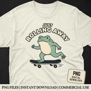Pode incluir: Camiseta creme com um desenho animado de um sapo andando de skate. O sapo é verde claro com a barriga creme e está suando. O texto "JUST ROLLING AWAY" está arqueado acima do sapo. Uma pequena etiqueta de download digital PNG está no canto inferior direito.