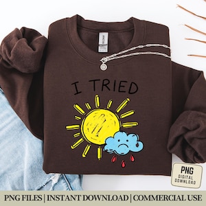 Könnte beinhalten: Ein braunes Sweatshirt mit dem Text "I Tried" über einer gelben Sonne und einer blauen Wolke mit roten Regentropfen. Eine silberne Halskette mit einem Sonnenanhänger befindet sich auf dem Sweatshirt. Das Bild enthält auch eine "PNG DIGITAL DOWNLOAD" Grafik.