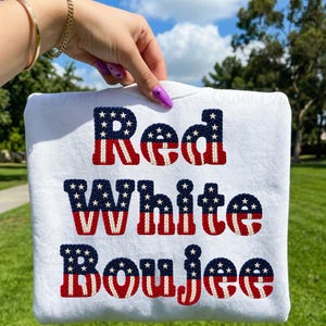 May include: White sweatshirt with the words "Red White Boujee" in a patriotic design. The letters are in red, white, and blue, resembling the American flag. The sweatshirt is held up against a background of green grass and a blue sky.