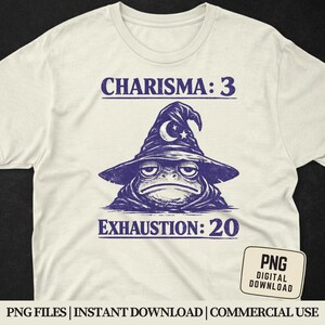 May include: Cream-colored t-shirt featuring a grumpy frog wearing a wizard hat with a crescent moon and star. The text reads "CHARISMA: 3" above the frog and "EXHAUSTION: 20" below. A small PNG digital download graphic is in the lower right corner.