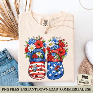 May include: Cream-colored Comfort Colors t-shirt featuring a graphic of two clogs decorated with red, white, and blue floral arrangements. One clog has a striped design, the other has stars. The shirt includes the text "PNG DIGITAL DOWNLOAD".