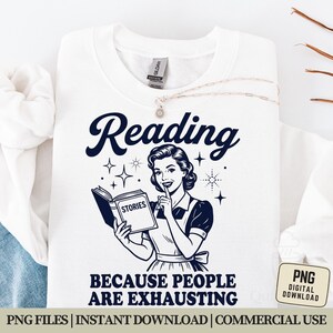 May include: White sweatshirt with the word "Reading" in navy blue script. Below the word is a vintage illustration of a woman holding a book labeled "Stories." The text "BECAUSE PEOPLE ARE EXHAUSTING" is below the illustration. A necklace and a tag are visible.