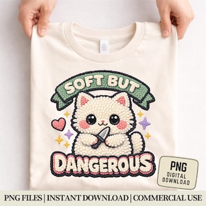 Op de afbeelding: Crèmekleurig T-shirt met een cartoonkat die een mes vasthoudt. Het ontwerp bevat de tekst "SOFT BUT DANGEROUS" boven en onder de kat, met een hart, sterren en een paarse vorm. Een digitale download tag is zichtbaar.