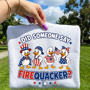 May include: White sweatshirt with a patriotic design featuring four cartoon ducks dressed in red, white, and blue attire. The text "DID SOMEONE SAY FIREQUACKER?" is printed on the front, along with stars.