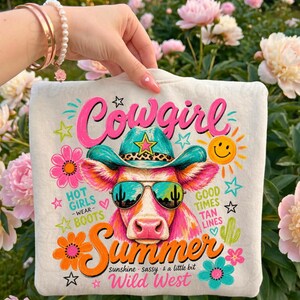 May include: Cream sweatshirt featuring a vibrant cow graphic wearing a turquoise cowboy hat and sunglasses. The design includes the word "Cowgirl" in pink and "Summer" in orange. Additional text reads "Hot Girls Wear Boots" and "Good Times Tan Lines."