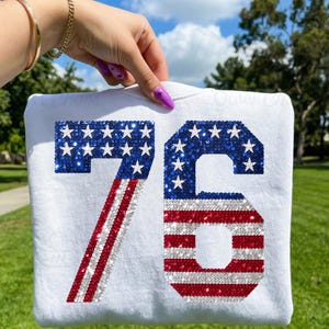 May include: White sweatshirt featuring the number 76 in a sequined design. The number 7 is red and white striped, while the 6 and the stars are blue with white stars, resembling the American flag.