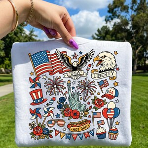 May include: White sweatshirt with a colorful patriotic design featuring the American flag, an eagle, the Statue of Liberty, fireworks, and the text "USA" and "Liberty". Includes a hot dog, cupcakes, and balloons. Perfect for celebrating the 4th of July.