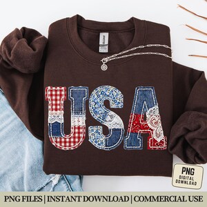 Op de afbeelding: Een bruine sweatshirt met het woord "USA" op de voorkant. De letters zijn gemaakt van patchworkstof in rood geruit, blauw bloemenpatroon en rood paisley-patroon. Een zilveren ketting met een hanger ligt op de sweatshirt.