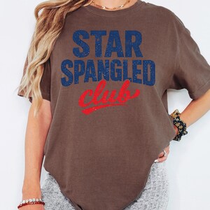 Può includere: T-shirt marrone con la scritta "STAR SPANGLED Club" in blu e rosso. La maglietta ha un aspetto vintage e usurato.