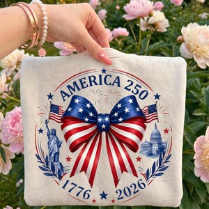 May include: Cream-coloured sweatshirt with a patriotic design. The design features a large bow in red, white, and blue, with the text "AMERICA 250" and the years "1776" and "2026". Includes images of the Statue of Liberty, the Capitol Building, and American flags.