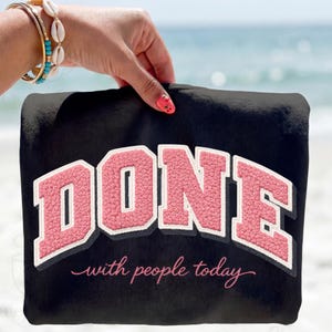 Op de afbeelding: Zwart sweatshirt met het woord "DONE" in roze, omlijnd met wit, en de zin "with people today" in cursief. Het sweatshirt wordt vastgehouden door een hand met een armband en nagellak.