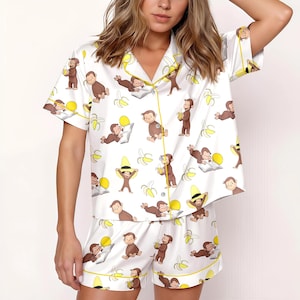 May include: White pajama set with a repeating pattern of a brown monkey, bananas, and yellow suns. The short-sleeved top and shorts have yellow trim. The pajama set is made of a soft, silky material.
