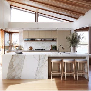 Puede incluir: Una cocina moderna con una isla de mármol y suelo de madera clara. La cocina cuenta con gabinetes de color claro, un techo de madera y grandes ventanas. Tres taburetes de madera con asientos grises están colocados en la isla.