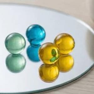 Pode incluir: Cinco bolas de gude de vidro em tons de azul, verde e amarelo estão sobre uma superfície refletora. As bolas de gude são redondas e transparentes, com detalhes internos visíveis. A superfície reflete as bolas de gude, criando imagens espelhadas.