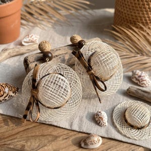 Op de afbeelding: Close-up van twee geweven oorbellen met een rond, open ontwerp. Elke oorbel heeft een bruine houten kraal en een decoratieve strik met een luipaardprint. De oorbellen worden getoond met schelpen en drijfhout.