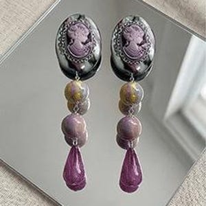 May include: A pair of dangle earrings with a cameo design in shades of purple and silver. The earrings feature a series of beads in varying shapes and colors, culminating in a teardrop-shaped charm. The earrings are displayed on a reflective surface.