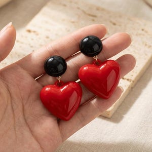 May include: A pair of heart-shaped earrings. Each earring features a glossy red heart pendant suspended from a black circular stud. The earrings are a bold statement piece, perfect for adding a pop of color to any outfit.
