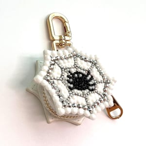 May include: A white, star-shaped keychain featuring a beaded spiderweb design with a central black spider. The web is accented with silver-toned beads and clear crystals. A gold-toned metal lobster clasp is attached to the top of the accessory.