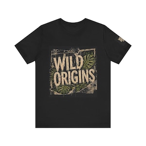 Könnte beinhalten: Schwarzes T-Shirt mit dem Text "WILD ORIGINS" in einem verwitterten Vintage-Stil. Das Design beinhaltet grüne tropische Blätter in einem rechteckigen Rahmen und erzeugt eine von der Natur inspirierte Grafik. Das T-Shirt hat kurze Ärmel und einen Rundhalsausschnitt.