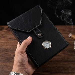 Puede incluir: Estuche de cigarros de cuero negro con costuras blancas y cierre plateado. El estuche cuenta con un higrómetro incorporado con un bisel dorado. El estuche está en la mano de una persona.