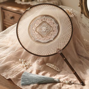 Può includere: Un ventaglio rotondo con cornice scura e centro in rete beige trasparente con intricati ricami bianchi e un motivo floreale centrale. Una nappa azzurra pende dal manico, accompagnata da due forcine di perle e un pettine di perle.