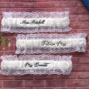 May include: Three white satin and lace garters with black embroidered text. The top garter reads "Mrs. Mitchell", the middle reads "Future Mrs", and the bottom reads "Mrs. Bennett". The garters are displayed on a brick wall.