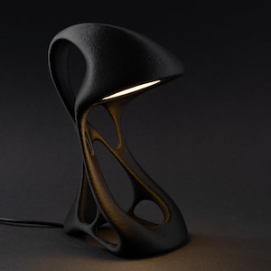 Op de afbeelding: Een zwarte tafellamp met een getextureerde, organische en vloeiende structuur met een gebogen, overhangende kap. De lamp heeft een open, onderling verbonden basisontwerp en straalt warm licht uit onder het bovenste gedeelte.